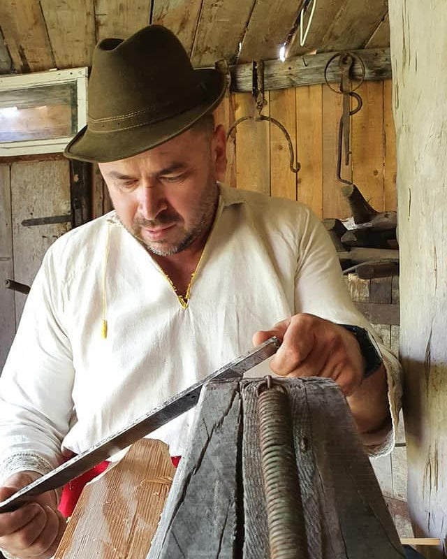 Meșter în costum tradițional cioplind manual o șindrilă de lemn prinsă într-un scaun de tâmplărie, în atelier